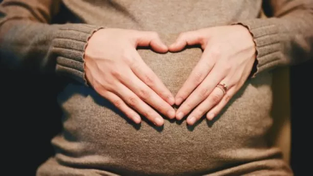 Frau in braunem Strickkleid, die sich herzförmig die Hände über den Schwangerschaftsbauch hält.